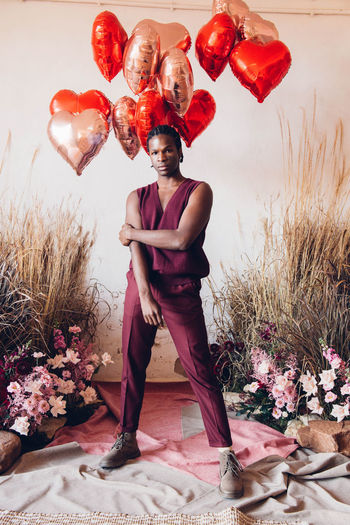 Full body of confident african american male in stylish outfit looking at camera while standing in room with flowers and heart shaped balloons