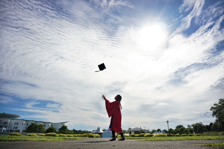 Side view of university student throwing mortarboard against sky during graduation