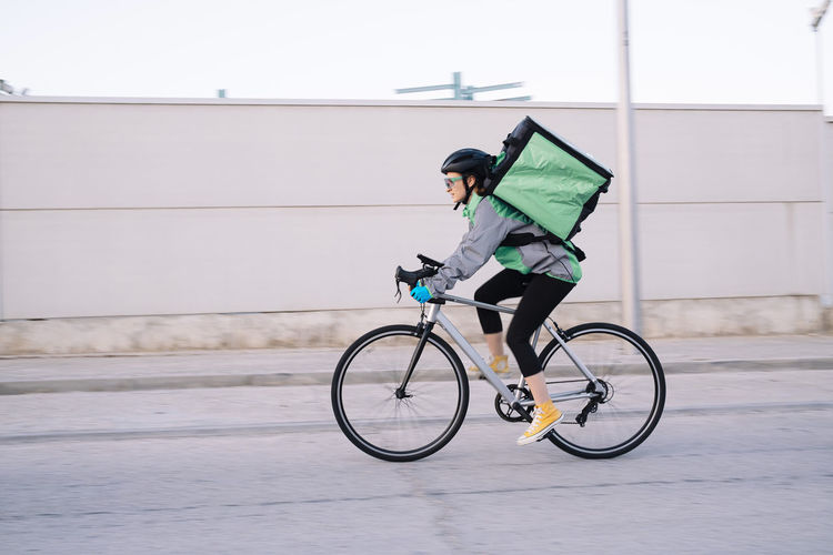 Side view of female courier with thermal bag riding bike on street road while delivering food in city, motion blur