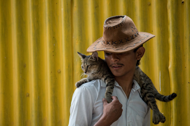 Teenage boy wearing hat with cat on shoulder against corrugated iron