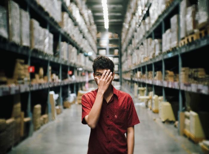 Man covering face with hand while standing amidst shelves at factory