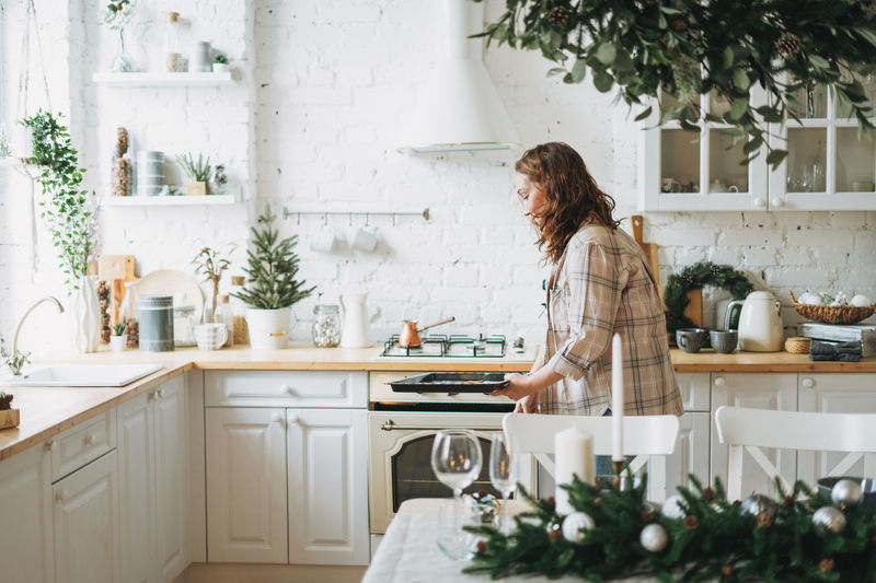 Attractive smiling woman with curly hair in plaid shirt bakes cookies at bright kitchen at home