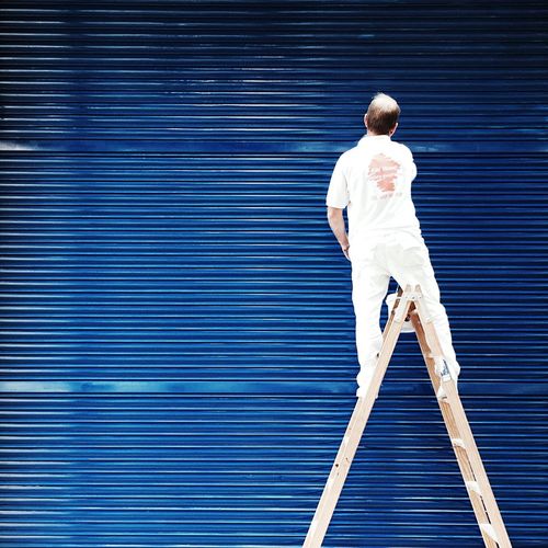 Rear view of man standing on ladder against blue corrugated iron