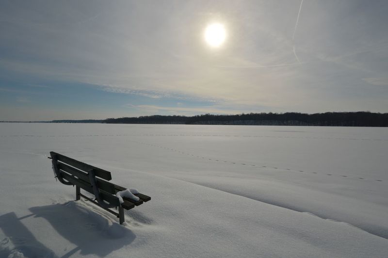 Bench on snow covered landscape against sunny sky