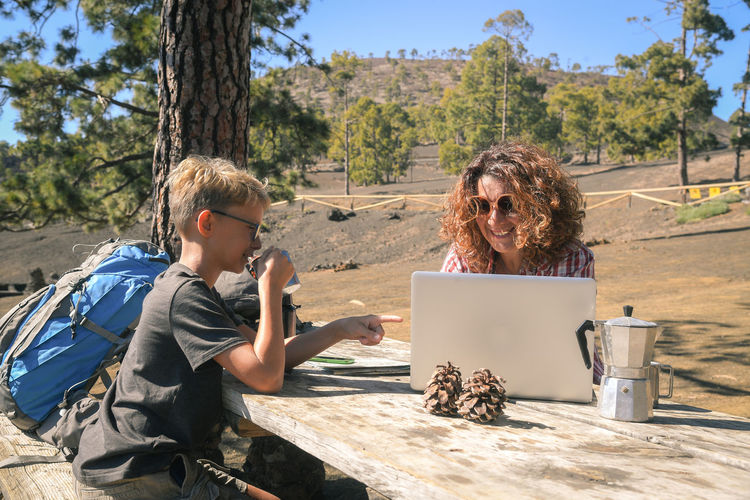 Smiling boy with coffee pointing towards mother using laptop on table