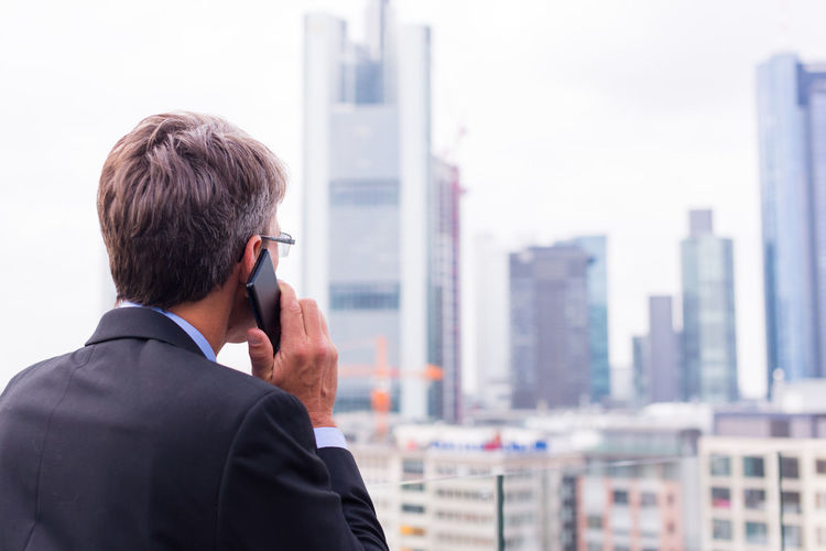 Mature businessman looking away while talking on smart phone against buildings in city