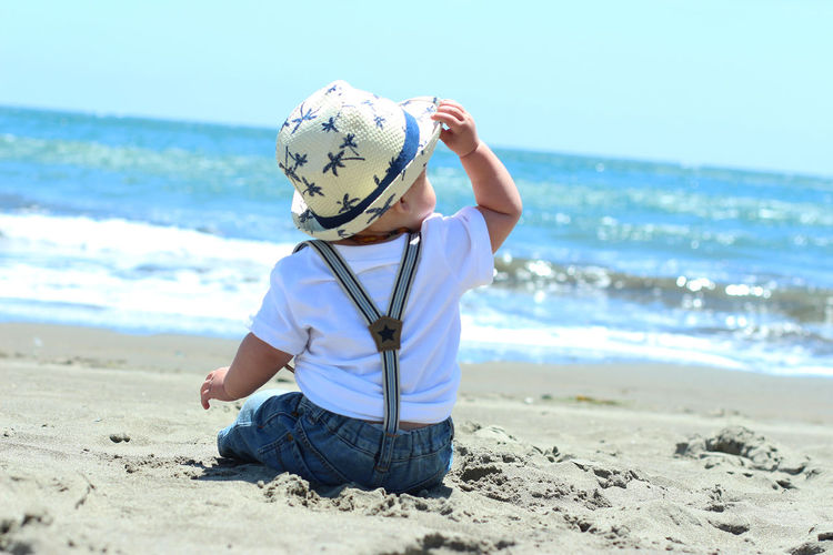 Rear view of fashionable baby in hat sitting on sea shore
