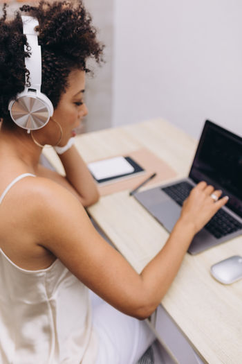 Black woman working on a laptop with headphones on, over shoulder view