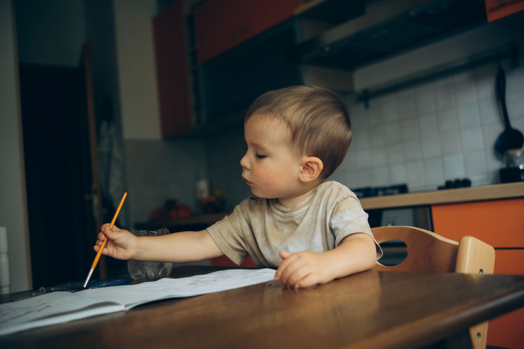 Child draws with a brush and paint in an album sitting at the table