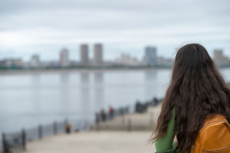 Girl with a backpack stands on the embankment of the river and admires the city on the other side.