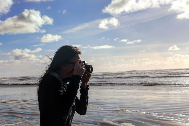 Woman photographing while standing on beach against sky