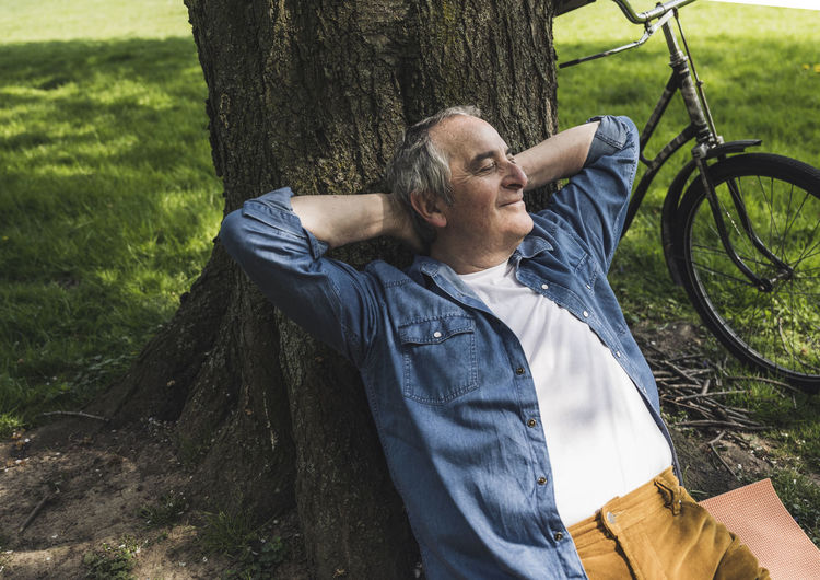 Smiling senior man leaning on tree trunk by bicycle at park