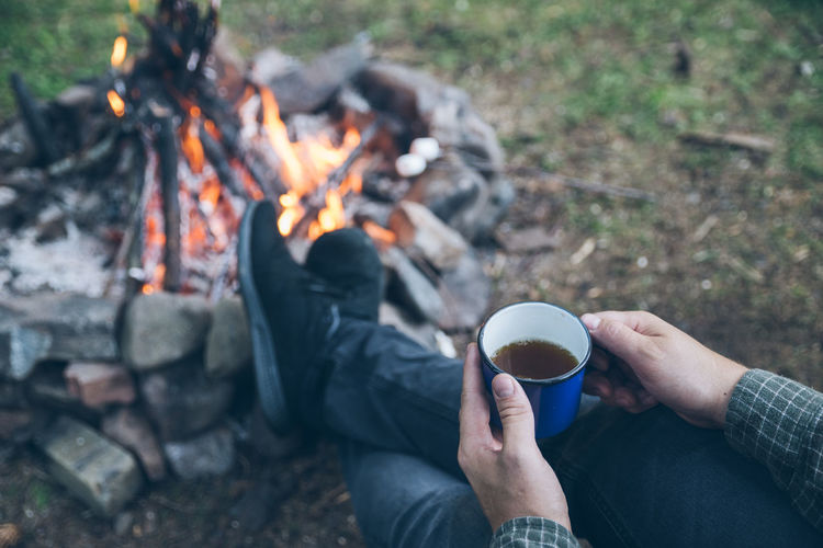 First point of view. man drinking tea in metal cup near camp fire. hiking