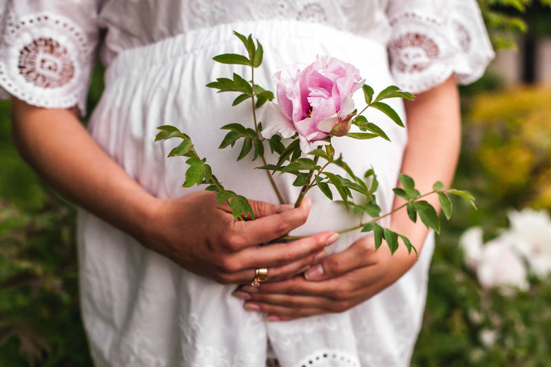 Midsection of pregnant bride holding pink flower while standing outdoors