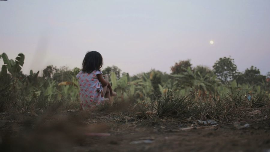 Rear view of girl sitting on field against sky at farm during sunset