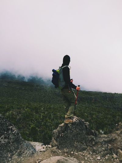 Side view of hiker standing on rock by forest against sky during foggy weather