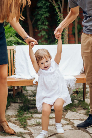 Little blonde girl playing jumping and holding her young parents hand in the garden. 