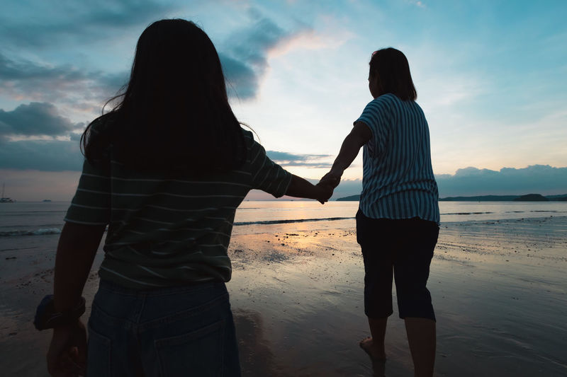 Rear view of mother and daughter holding hands while walking at beach against sky