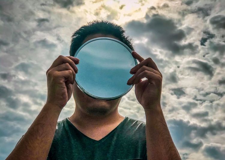 Portrait of man holding mirror against cloudy sky.