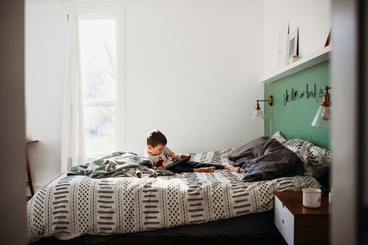Young boy laying alone in bed using tablet in modern bedroom