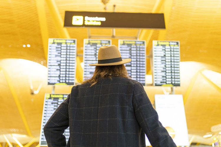 From below back view of stylish male traveler in hat checking information about flight on timetable display in airport