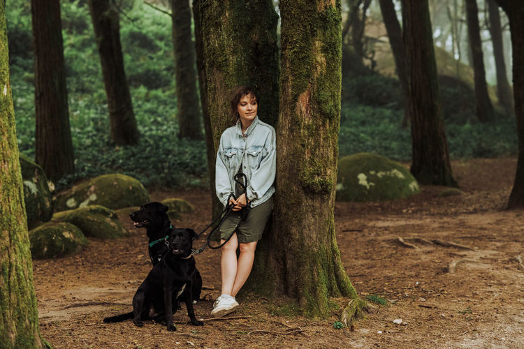 Woman with two dogs in the forest leaning against a mossy tree