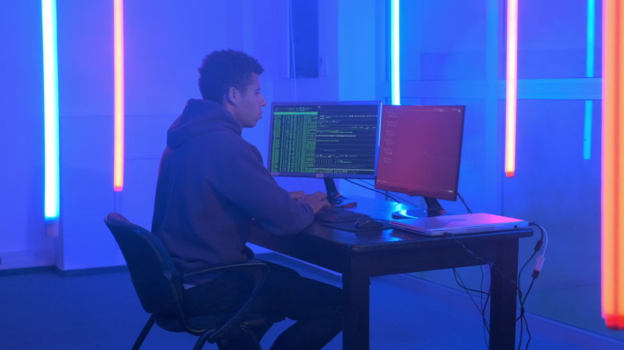 Young man in hood hacking by using computer while sitting at illuminated office