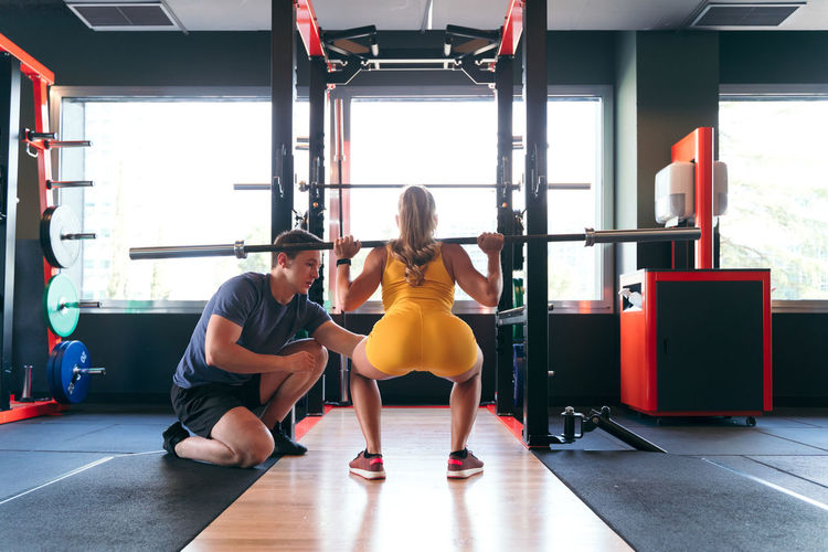 Back view of unrecognizable female athlete doing squats with heavy barbell during training in gym under supervision of male personal trainer