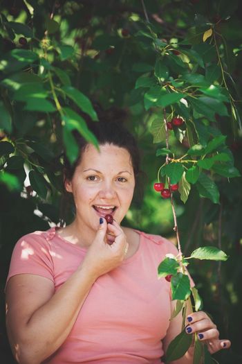 Portrait of smiling mid adult woman eating cherry by tree