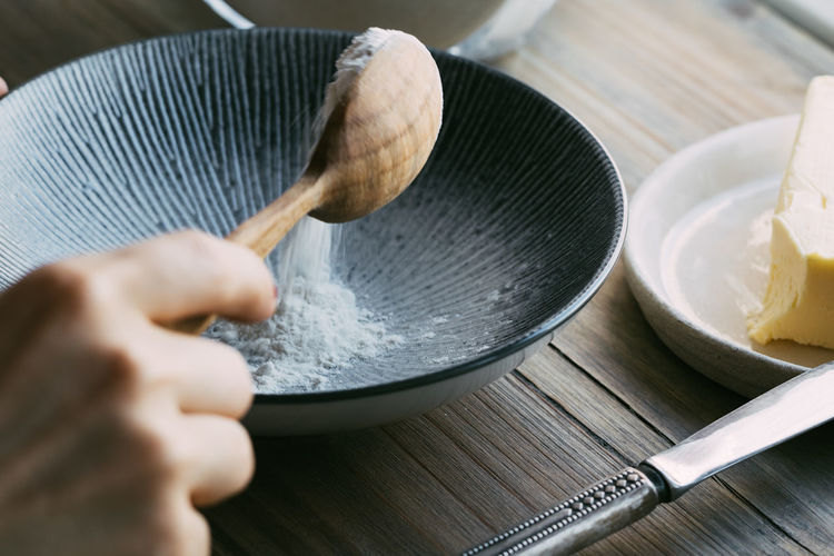 Cropped image of woman hand putting flour with wooden spoon in bowl at table