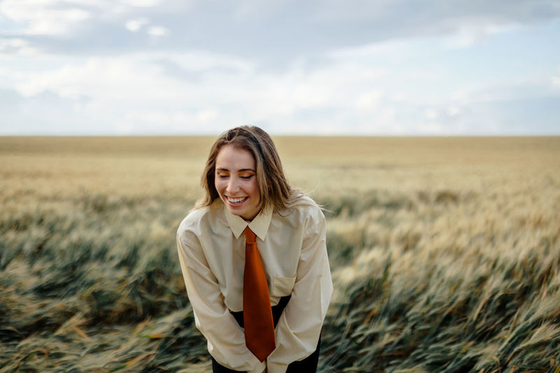 Positive young female in white shirt and red tie standing with hands behind back among wheat spikes in countryside