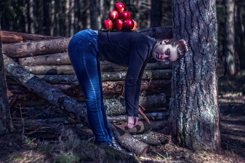 Full length of woman bending with apples on back by tree trunk in forest