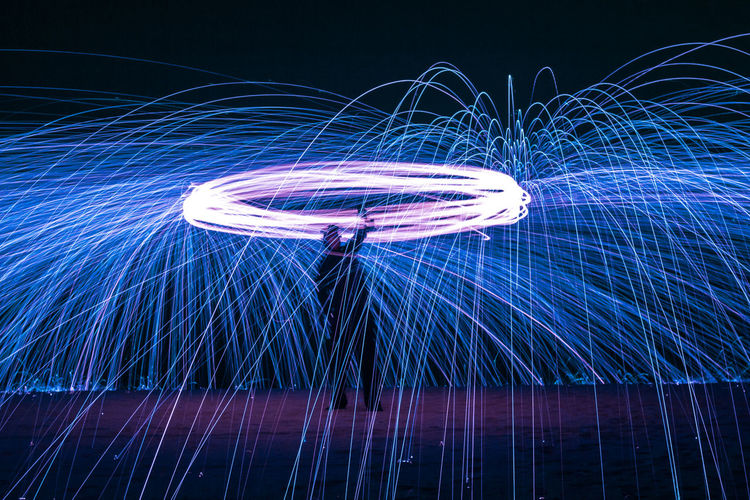 Long exposure silhouette of unrecognizable person spinning burning stick emitting sparkles while dancing with fire at night