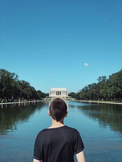 Rear view of boy looking at lake in city against blue sky
