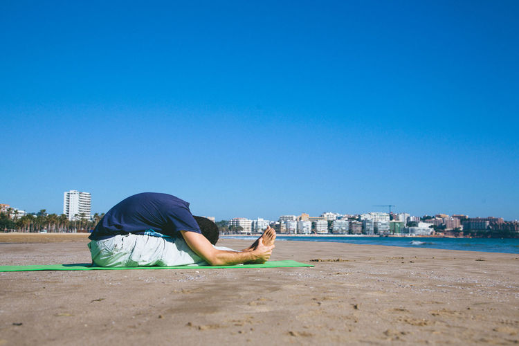 Man stretching on beach against clear blue sky