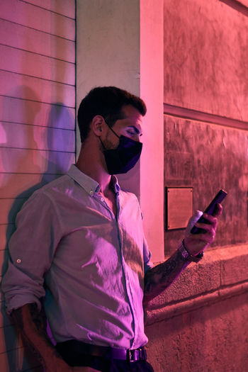 Young man with a mask looks at his mobile phone at night
