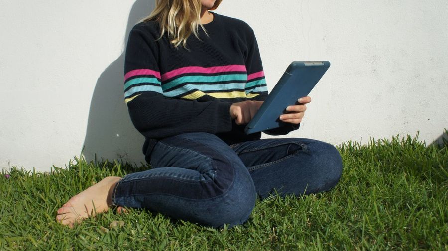 Low section of woman using digital tablet while sitting on field against wall