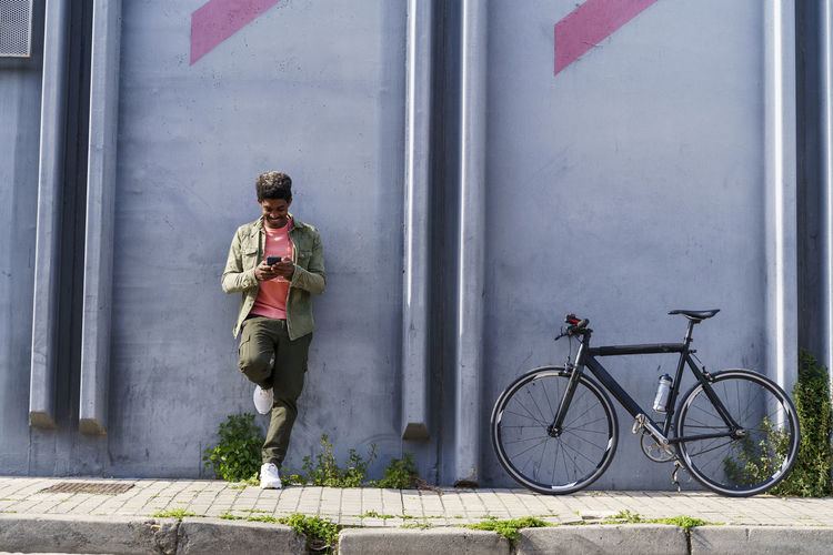 Smiling man with bicycle using smart phone while leaning on wall at footpath