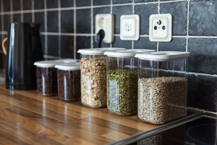 Plastic containers with muesli and sunflower and pumpkin seeds placed near coffee beans and electric kettle on counter in kitchen