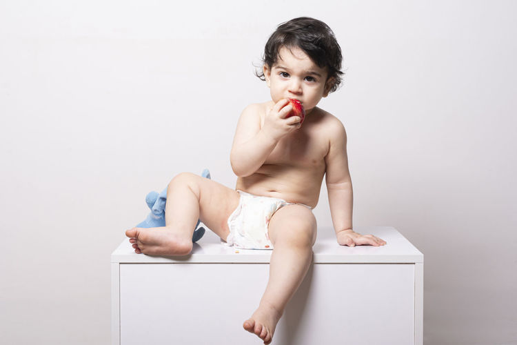 One year old baby wear diaper and eating an apple on a white table