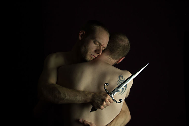 Close-up of shirtless gay couple embracing against black background