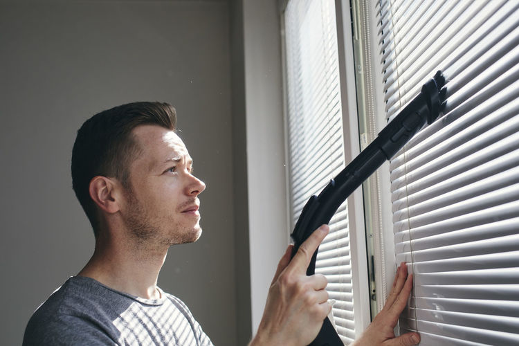 Man cleaning dust from window blind by vacuum cleaner at home. themes housework and housekeeping.