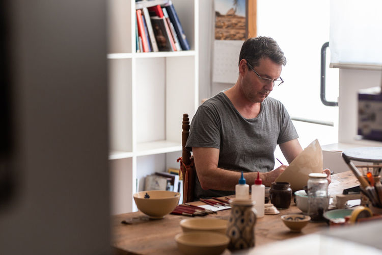 Man sitting at table with brushes and drawing sketches on handmade ceramic plate