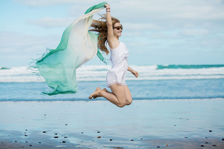 Woman holding shawl while jumping on shore at beach