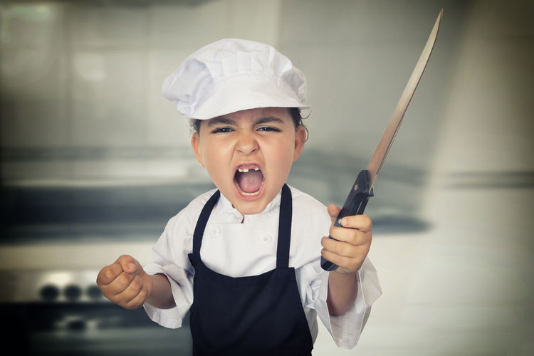 Portrait of girl wearing chef uniform holding knife while screaming in kitchen