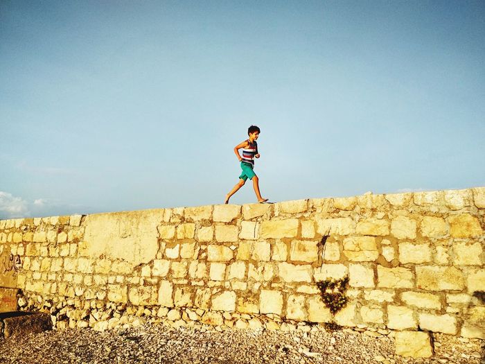 Side view of boy walking on retaining wall against blue sky