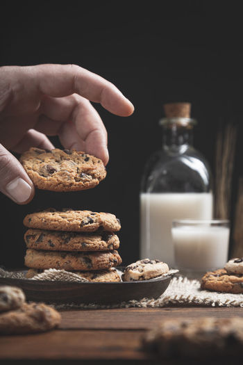 Hand holding chocolate chip cookie with glass and a bottle of milk on wooden base, dark background,.