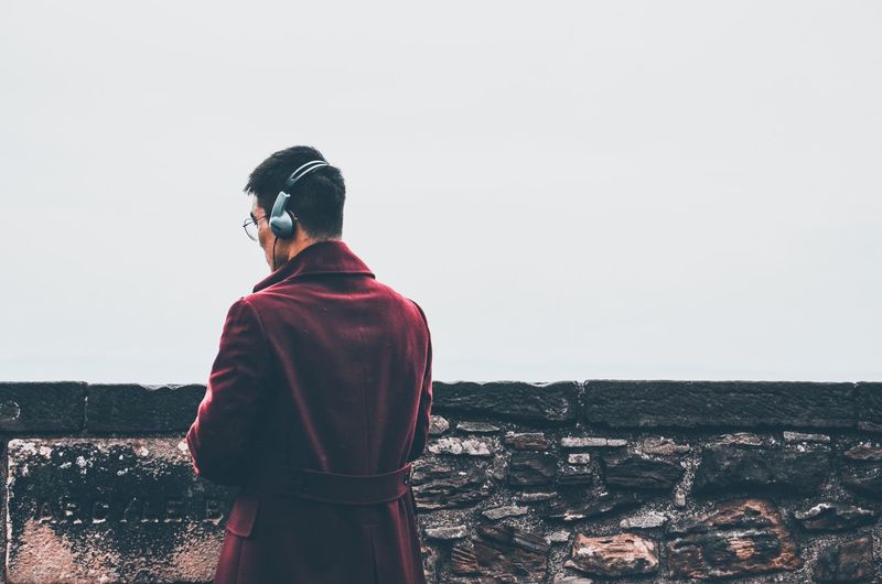Rear view of man listening music while standing by retaining wall against clear sky