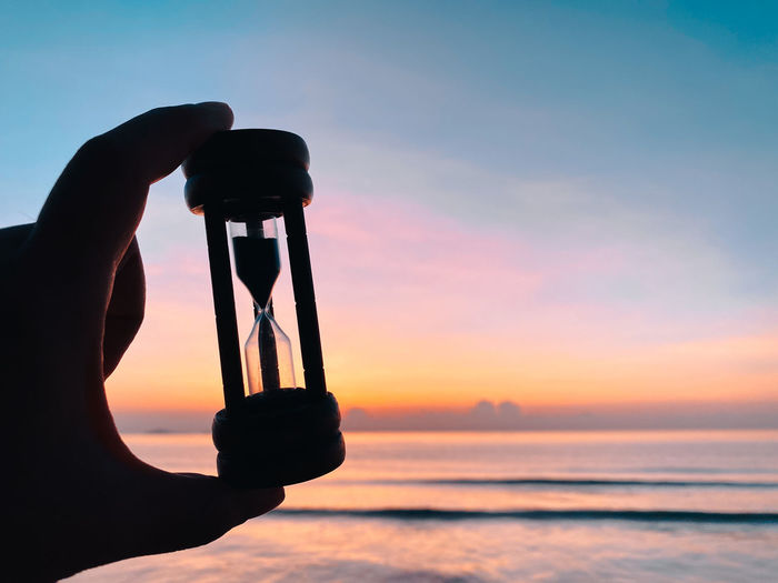 Person holding hourglass against sea against sky during sunset