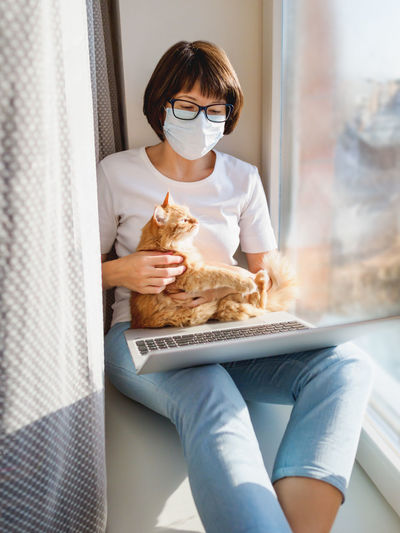 Woman in medical mask remote works with laptop and cute ginger cat. quarantine coronavirus covid19.